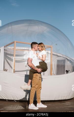 Vista laterale dell'appassionato uomo che tiene e abbraccia la giovane donna mentre si trova di fronte a un romantico glamping trasparente Foto Stock