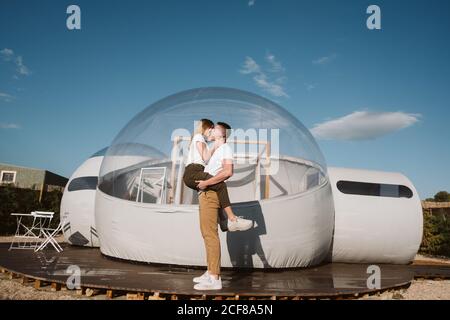 Vista laterale dell'appassionato uomo che tiene e abbraccia la giovane donna mentre si trova di fronte a un romantico glamping trasparente Foto Stock