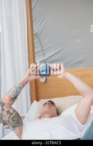 Bell'uomo tatuato in una maglietta bianca che si ritorce in mani piccole palla di terra mentre si rilassa a letto Foto Stock