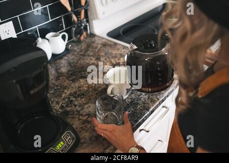 Ritagliare la vista laterale di una donna irriconoscibile nel cappello con orologio da polso versare il caffè appena preparato in tazze bianche mentre si è in piedi cucina a casa Foto Stock