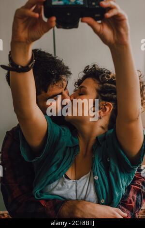Affettuosa coppia hipster baciando e prendendo selfie con foto sopra macchina fotografica mentre si è seduti sul pavimento a casa Foto Stock