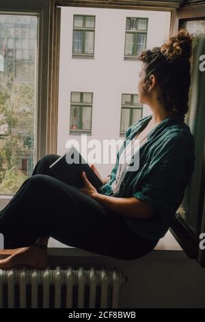 Vista laterale di una donna rilassata in vestito casual in possesso di un libro mentre si siede a piedi nudi sul davanzale della finestra in appartamento guardando fuori dalla finestra Foto Stock