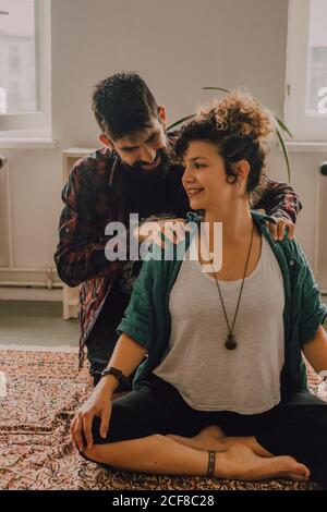 Uomo in vestito casual massaggiante schiena di rilassato Donna seduta in posizione lotus sul pavimento a casa Foto Stock