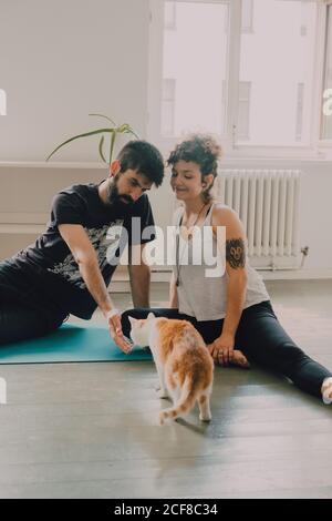 Vista laterale della coppia tenera che si abbraccia mentre si siede sul pavimento accanto al gatto zenzero a casa Foto Stock