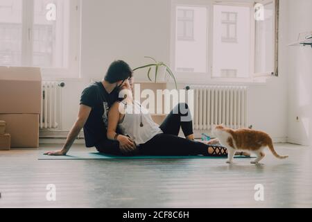 Vista laterale della coppia tenera che si abbraccia mentre si siede sul pavimento accanto al gatto zenzero a casa Foto Stock