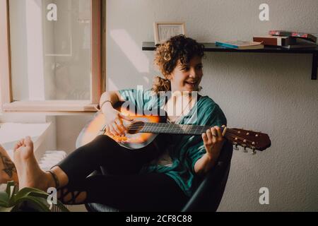 Donna a piedi nudi rilassati in abiti casual suonando chitarra acustica mentre si siede sulla sedia accanto alla finestra a casa Foto Stock