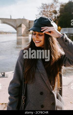 Felice giovane donna in outerwear alla moda sorridente a occhi chiusi e di regolare il tappo mentre si sta in piedi sulla banchina vicino al fiume giorno di sole Foto Stock