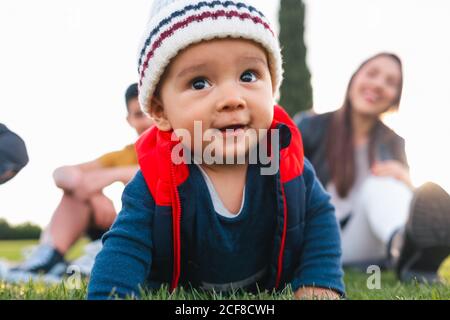 Cute bambino etnico in abiti caldi sorridendo mentre strisciando sopra prato verde durante il fine settimana in famiglia in campagna Foto Stock