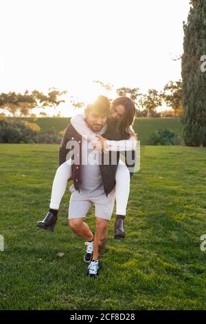 Giovane uomo allegro che dà giro piggyback alla moglie felice mentre godetevi una soleggiata giornata autunnale su un prato verde in campagna Foto Stock