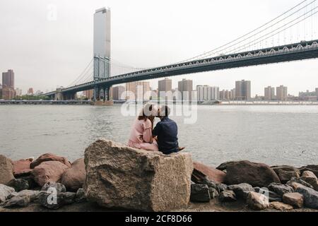 Vista laterale di una coppia amorevole che abbrancava e baciava mentre si è seduti Sulle pietre vicino al fiume sullo sfondo del ponte di Brooklyn durante passeggiata in città Foto Stock