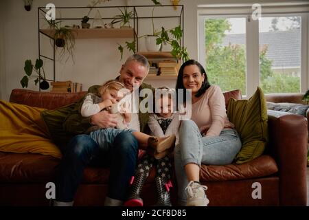 Coppia allegra seduta sul divano con due belle piccole figlie a casa - un grande felice ritratto di famiglia su uno divano moderno a casa Foto Stock