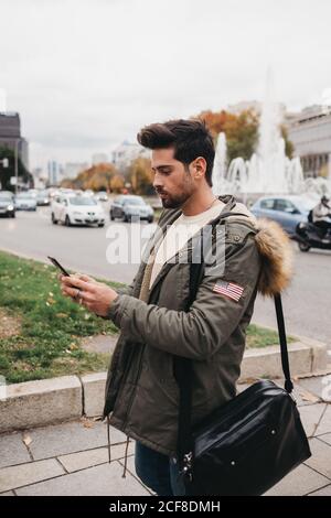 Vista laterale dell'uomo alla moda in parka in piedi con mobile telefono e ricerca su sfondo urbano Foto Stock