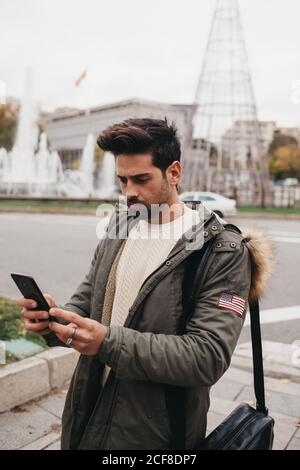 Vista laterale dell'uomo alla moda in parka in piedi con mobile telefono e ricerca su sfondo urbano Foto Stock