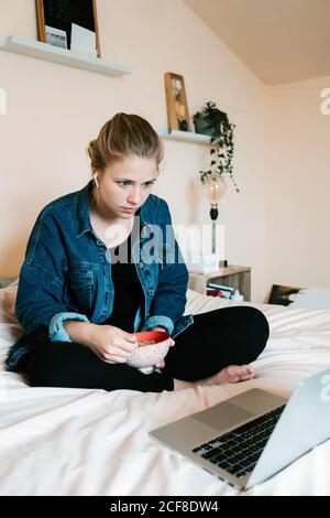 Donna a piedi nudi con stordito e pusivo in veri auricolari wireless e casual vestiti con ciotola di cibo gustoso che si concentra sullo schermo con interesse mentre si siede con le gambe incrociate sul letto morbido e. utilizzo di netbook in un accogliente appartamento Foto Stock