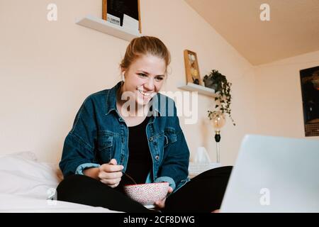 Donna a piedi nudi con stordito e pusivo in veri auricolari wireless e casual vestiti con ciotola di cibo gustoso che si concentra sullo schermo con interesse mentre si siede con le gambe incrociate sul letto morbido e. utilizzo di netbook in un accogliente appartamento Foto Stock