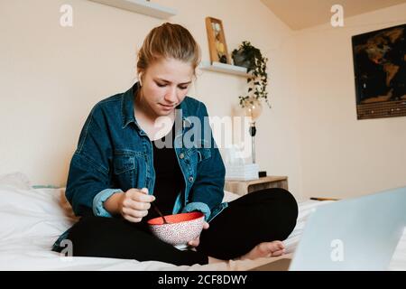 Donna a piedi nudi con stordito e pusivo in veri auricolari wireless e casual vestiti con ciotola di cibo gustoso che si concentra sullo schermo con interesse mentre si siede con le gambe incrociate sul letto morbido e. utilizzo di netbook in un accogliente appartamento Foto Stock