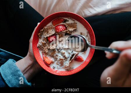 Da sopra raccolto femmina in abiti casual che tengono cucchiaio di metallo e ciotola rossa con pezzi di fragola e farina d'avena versato con latte mentre si siede sul letto nella camera da letto luminosa Foto Stock