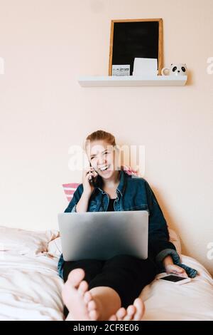 Positivo rilassato giovane femmina in abiti casual guardando via e. ridendo mentre si siede con le gambe incrociate con il computer portatile su morbido letto e telefonate in una camera accogliente Foto Stock