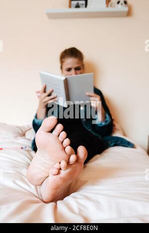 Blurred nuefoot femmina in vestiti casual leggere libro con interesse mentre vi rilassate da soli su un letto morbido, vi senterete sicuri a casa Foto Stock