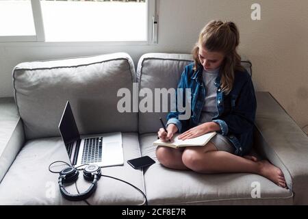 Alto angolo di una giovane donna seria e mirata in un abbigliamento casual prendere appunti in diario mentre si siede sul divano con gadget in soggiorno Foto Stock