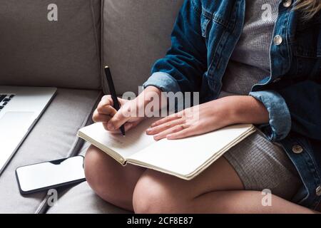 Crop giovane femmina in abito casual prendendo appunti in diario mentre si siede sul divano con gadget in soggiorno Foto Stock