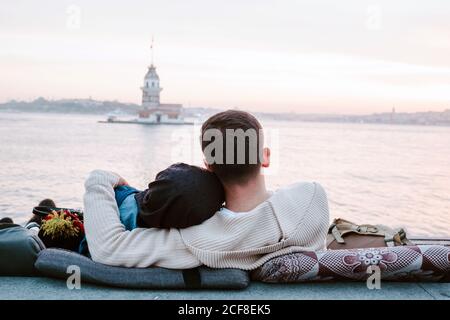 Vista posteriore di coppia anonima amorevole abbracciando seduta su cuscini sul lungomare e godendo del tramonto meraviglioso durante la data romantica in Istanbul Foto Stock
