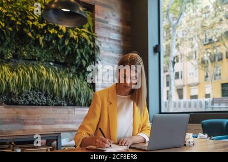 Donna d'affari in abiti colorati scrivere con la penna in notebook mentre si siede al tavolo di legno e utilizzando il computer portatile in una caffetteria moderna e leggera Foto Stock