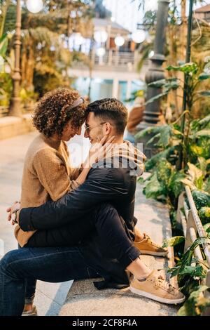 Vista laterale del giovane uomo e della donna che abbraccia e tocca la fronte mentre si siede sulla panchina durante la data romantica sulla strada della città Foto Stock