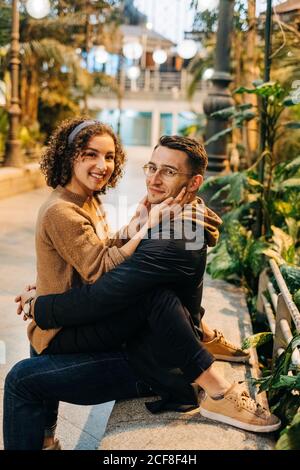 Vista laterale del giovane uomo e della donna che abbraccia e guarda la macchina fotografica mentre si siede sulla panca durante la data romantica sulla strada della città Foto Stock