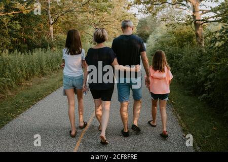 Vista posteriore di uomo e donna adulti con figlie vestite in t-shirt casual e pantaloncini a piedi sul sentiero tra alberi verdi, mentre trascorrere la giornata estiva insieme in campagna Foto Stock