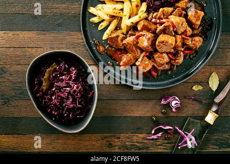 Manzo tedesco piccante o ragù di selvaggina con cavolo rosso E patatine fritte croccanti condite con peperoncino servito su a. tavolo rustico in una vista dall'alto verso il basso Foto Stock