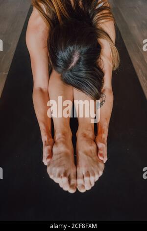 donna irriconoscibile barefooted in activewear stretching corpo mentre facendo paschimottanasana mettiti in posa sul tappeto sportivo al piano della moderna palestra Foto Stock