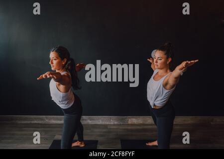 Le donne in abbigliamento sportivo facendo guerriero pongono due esercizi di yoga in piedi su tappetini sportivi nella moderna palestra Foto Stock