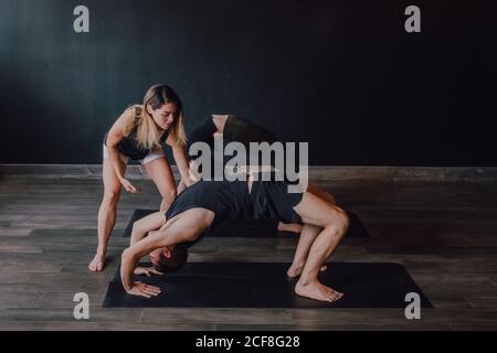 Alto angolo di adulto barefooted istruttore in abbigliamento sportivo che sostiene l'uomo mentre insegnano gruppo di persone sportive a fare urdhva dhanurasana posa in una moderna palestra Foto Stock