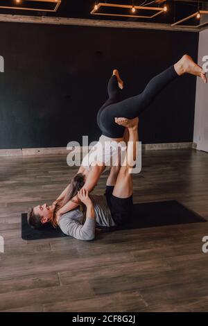 Vista laterale di un gioioso atletico con una giovane partner femminile pratica acro yoga insieme in foglia ripiegata posa su nero stuoia insieme in una moderna palestra scura Foto Stock