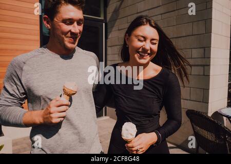 Crop uomo adulto e positivo FIT Donna con gli occhi chiusi in abiti casual divertirsi e ridere mentre si cammina lungo la strada e mangiare gelato yummy Foto Stock