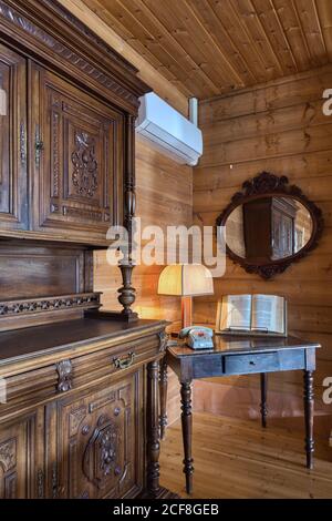 Tavolo con libro aperto con lampada e telefono in angolo di interni in vecchio stile con specchio su pareti di legno e armadio Foto Stock