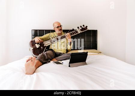 Uomo anziano seduto a letto con seduta e prendendo appunti in un notebook mentre guardi una lezione online sul tablet Foto Stock