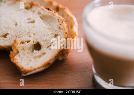 Preparare il caffè fresco con il latte in una tazza di vetro trasparente vicino a fette di pane fresco in cucina tavolo di legno Foto Stock