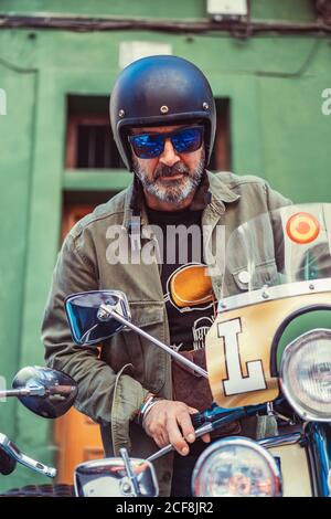 Uomo maturo bearded in casco e occhiali seduti su ordinazione moto con molti fari sulla strada della città Foto Stock