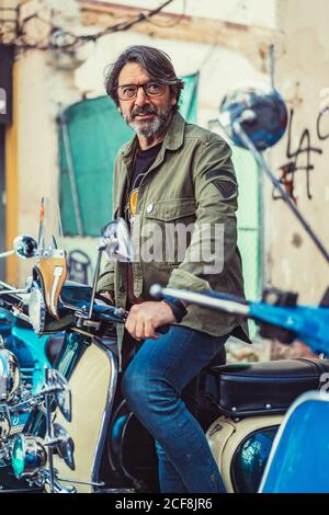 Uomo maturo bearded in occhiali che siedono su moto su ordinazione con molti fari sulla strada della città Foto Stock