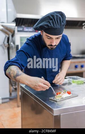 Concentrato professionista chef maschile guarnire e rifinire delizioso piatto pronto per servire mentre si lavora al banco metallico nella cucina del ristorante Foto Stock