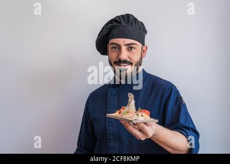 Felice cuoco adulto bearded in uniforme blu scuro e nero cappello che tiene in mano piatto con piatto gourmet e guardare sulla fotocamera mentre si è in piedi contro la parete bianca Foto Stock
