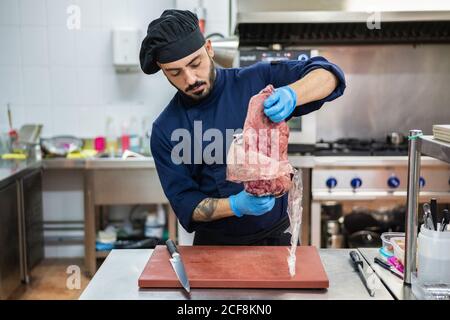 Chef maschile in uniforme blu scuro e guanti che tirano grandi pezzo di carne cruda fuori della confezione di plastica sottovuoto mentre in piedi davanti alla barra con taglierina e coltello in cucina Foto Stock