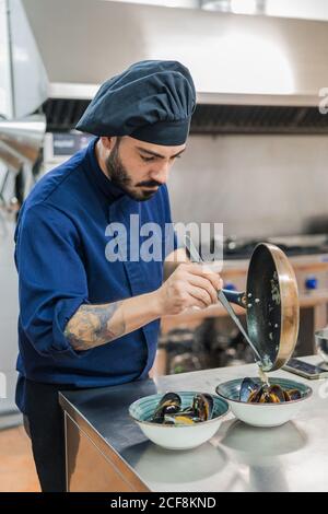 Cuoco maschile serio in cappello nero finishing piatto e mettere ingredienti dalla padella alle ciotole con cozze mentre si è in piedi al bancone in metallo in cucina professionale Foto Stock