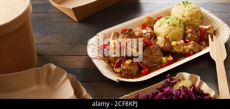 Servizio da asporto di goulash di manzo e gnocchi in un monouso piastra di cartone con forchetta di legno con una porzione laterale di cavolo rosso Foto Stock