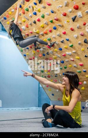 Vista laterale dell'atleta soddisfatta seduto in posa lotus sul pavimento mentre sorridi e guarda via durante l'allenamento di arrampicata in palestra Foto Stock