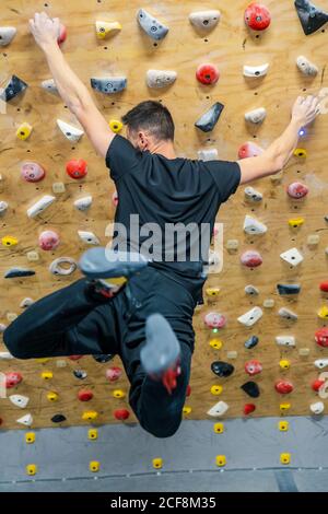 Vista posteriore di un giovane uomo con impugnature di tenuta attive stretto mentre appeso in aria durante l'allenamento di arrampicata in palestra Foto Stock