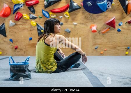 Vista posteriore della giovane atleta in abbigliamento sportivo su cui riposarsi pavimento e guardando via con parete di arrampicata colorata su sfocato sfondo in palestra Foto Stock