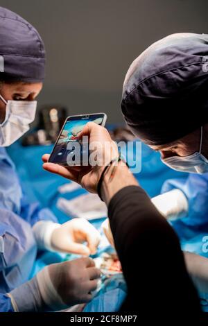 Ritaglio ad angolo elevato maschio che fotografa con smartphone mentre chirurghi concentrati in interventi chirurgici ad esecuzione uniforme utilizzando attrezzi speciali in sala operatoria di ospedale moderno Foto Stock